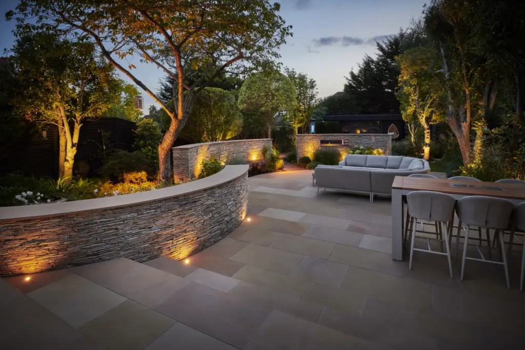 Buckhurst Hill at dusk: the curved paddlestone wall, uplit trees and flagstone terrace of the BALI award-winning garden, designed and built by Refined Landscapes