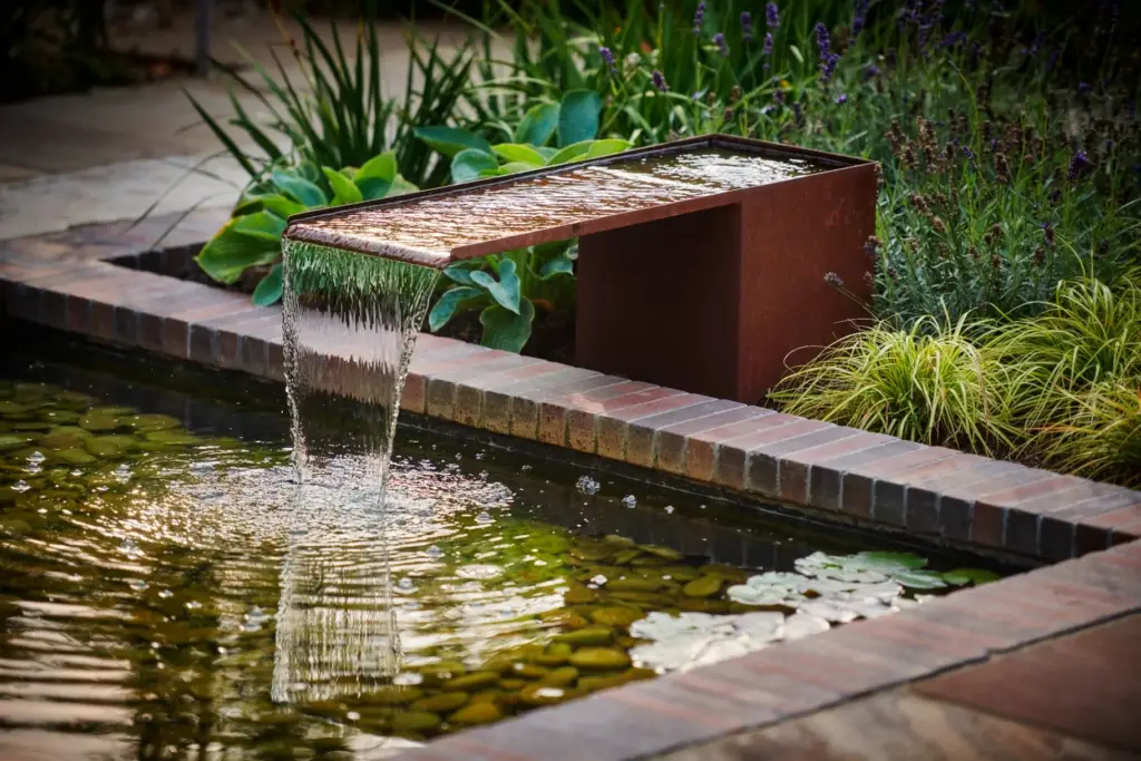 Corten steel water feature cascading into brick-edged pond with lily pads