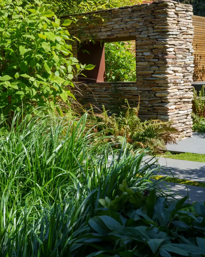 A drystone wall with a square opening, seen through grasses and a deep planted border