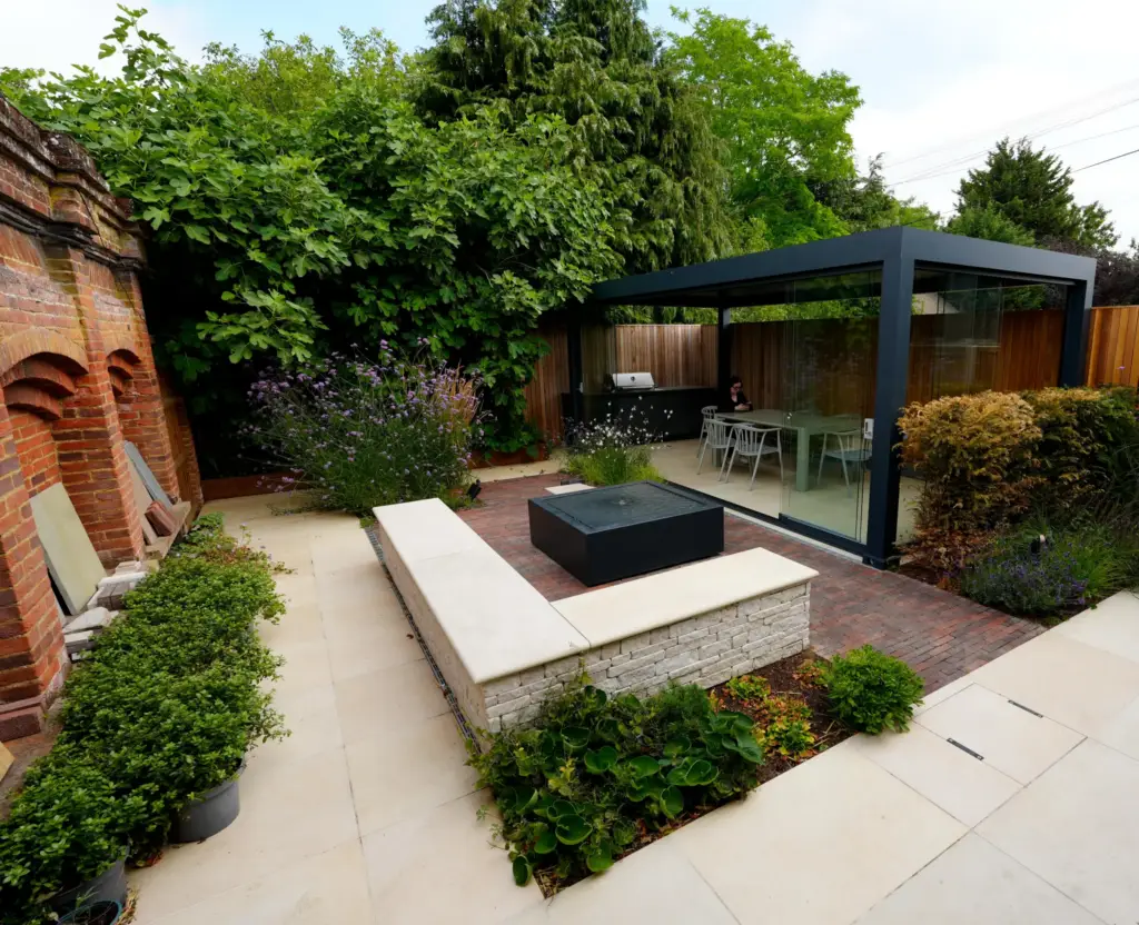 Inside the showroom at Ickleford Manor: a dining table beneath the louvred pergola, looking out to the courtyard, its cube water feature and the paving samples in the brick arches