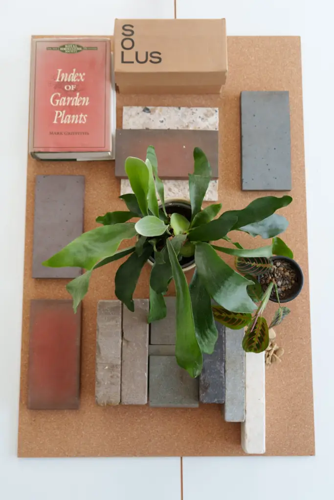 A material sample board laid out with stone, porcelain and brick samples, a planting reference book and two plants
