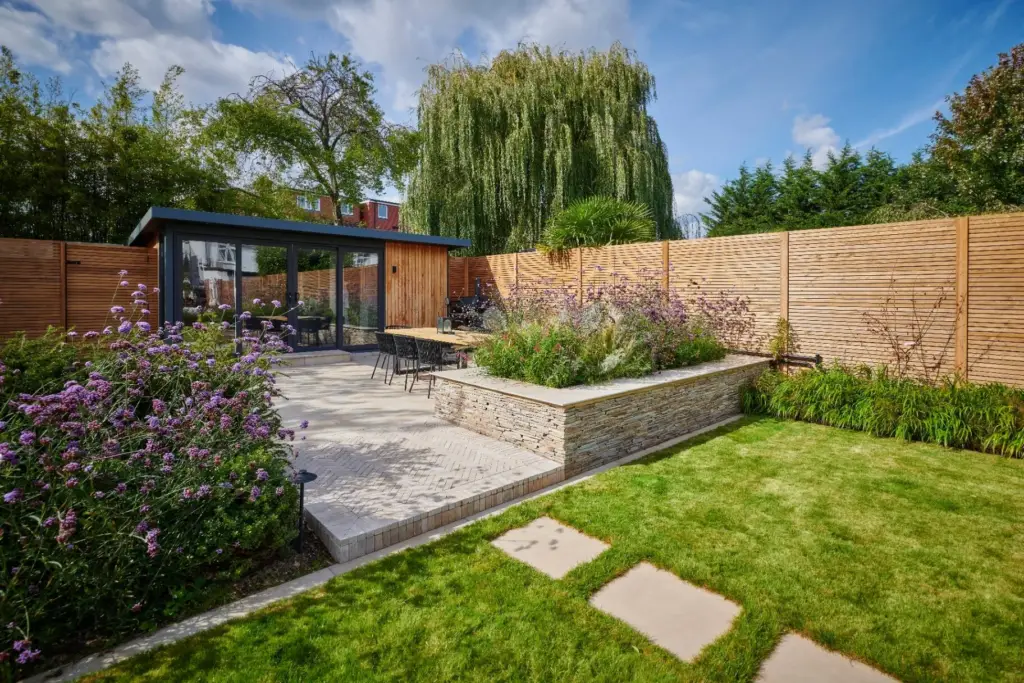 Contemporary garden with a paved terrace, raised stone planting beds, lawn and timber garden room.